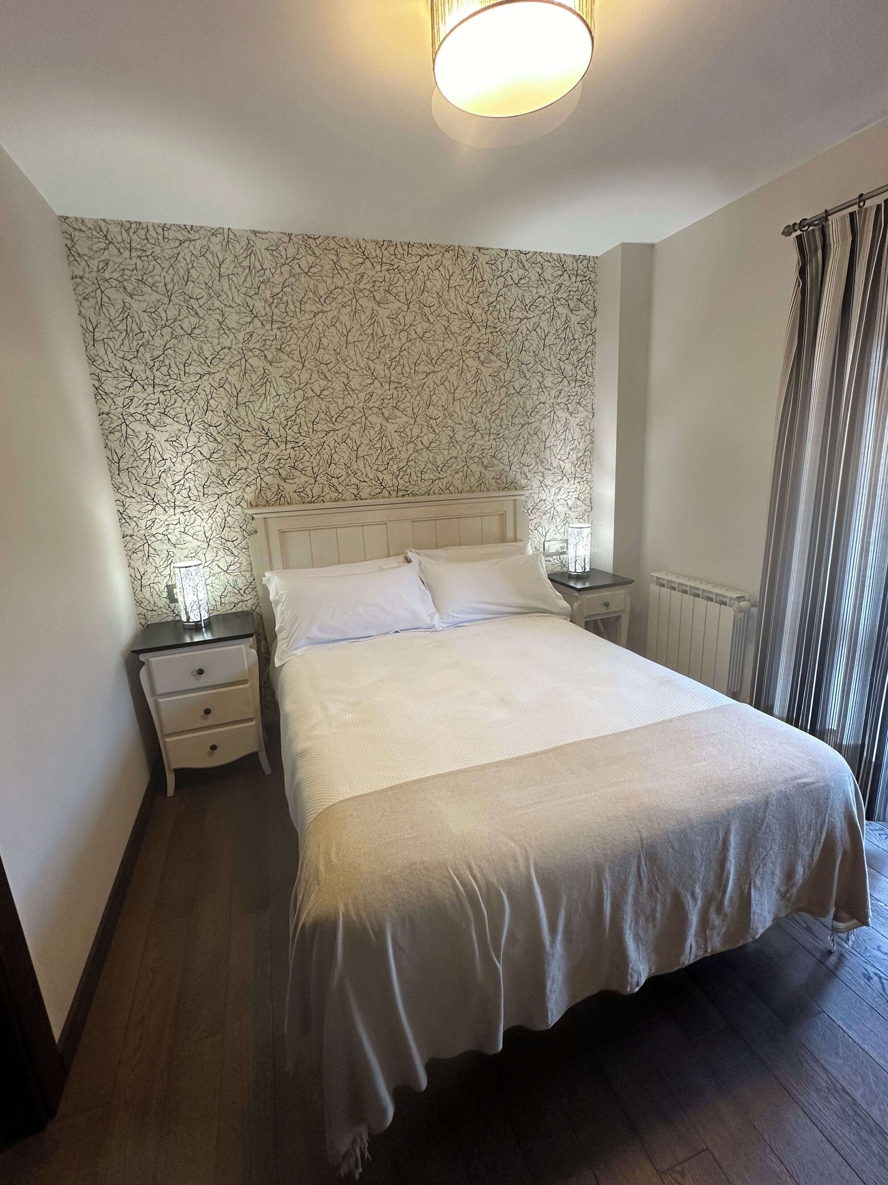 Un dormitorio con una cama grande, ropa de cama blanca, una mesita de noche con lámpara y un papel tapiz estampado.