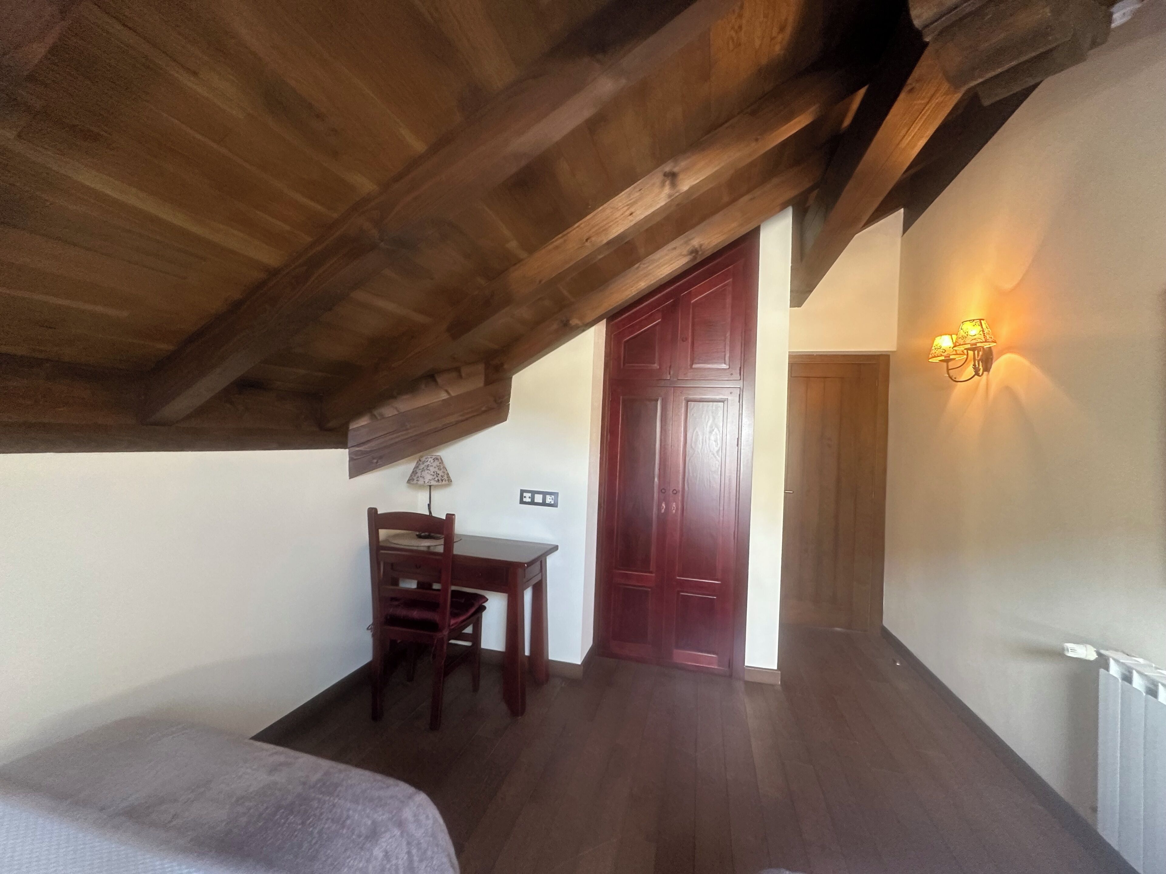 Quarto com teto de madeira inclinado, porta de madeira, uma pequena mesa com uma cadeira e luminária fixada na parede.