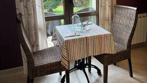 A dining area with a table set for two, wicker chairs, a striped tablecloth, and a view of greenery outside. - Hospedería Rural Santa Cruz (Socobio)