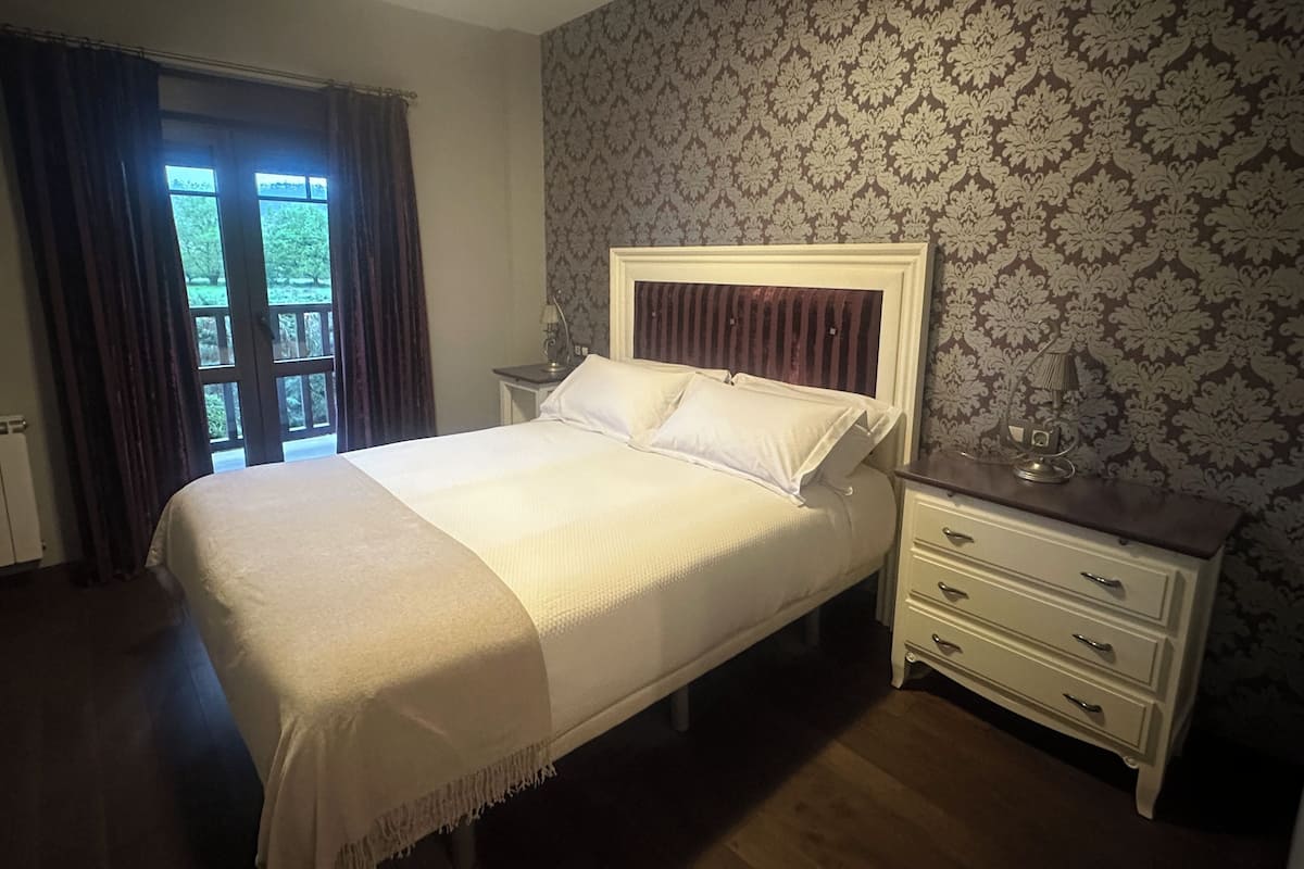 A neatly made bed with a headboard, a bedside table with a lamp, and a window with curtains.