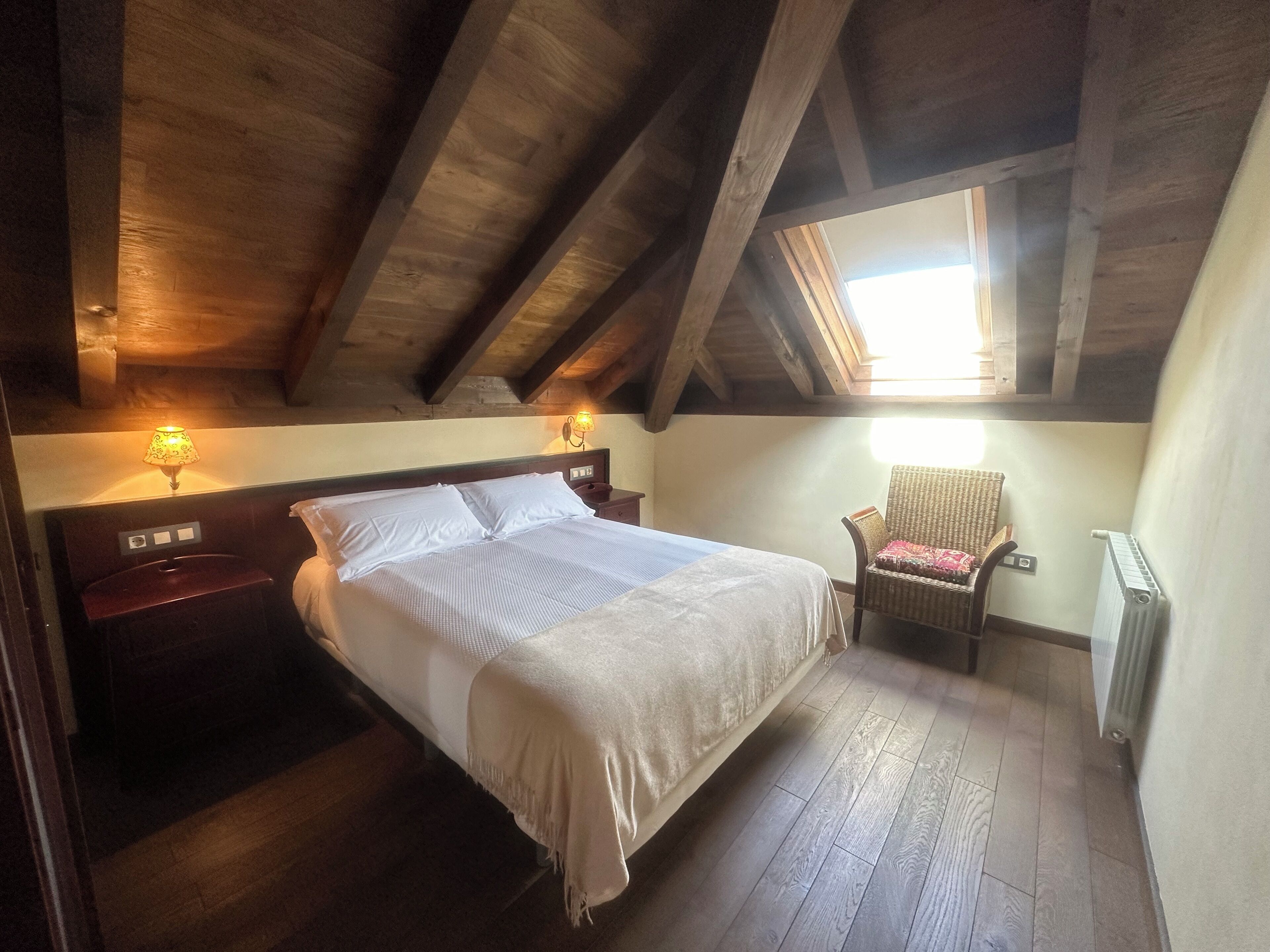 A bedroom with a large bed, wooden beams, a window, a chair, and a nightstand with a lamp.