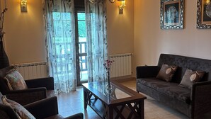 A living room with a dark sofa, two armchairs, a glass coffee table, a chandelier, and a wall-mounted light fixture. - Hospedería Rural Santa Cruz (Socobio)