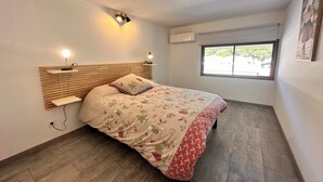 A bedroom with a wooden headboard, a bed with floral bedding, bedside lamps, a window with a view, and an air conditioning unit.
