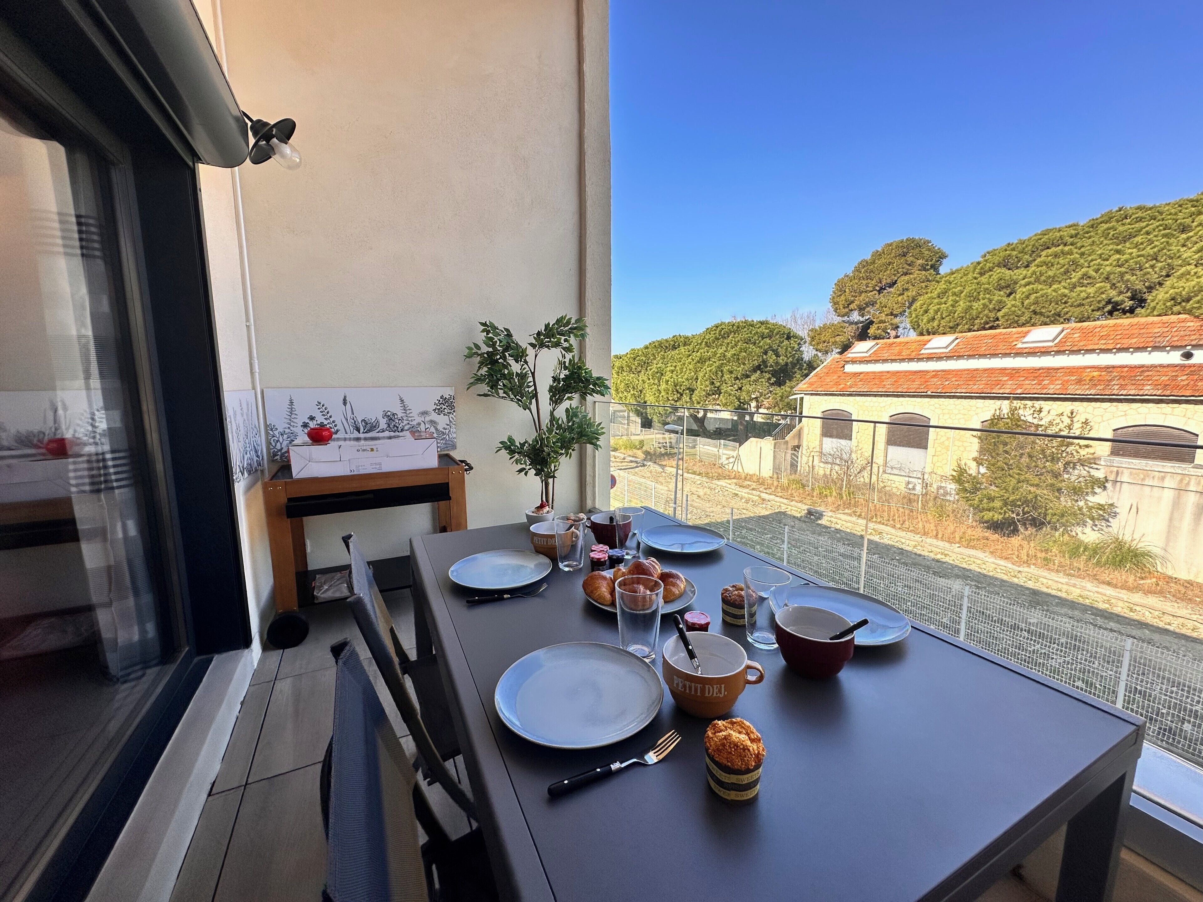 A balcony with a dining table set for two, a small plant, and a view of a building with a red roof.