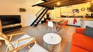 A modern living room with a TV, wicker chairs, a red sofa, and a dining area with a kitchen in the background.