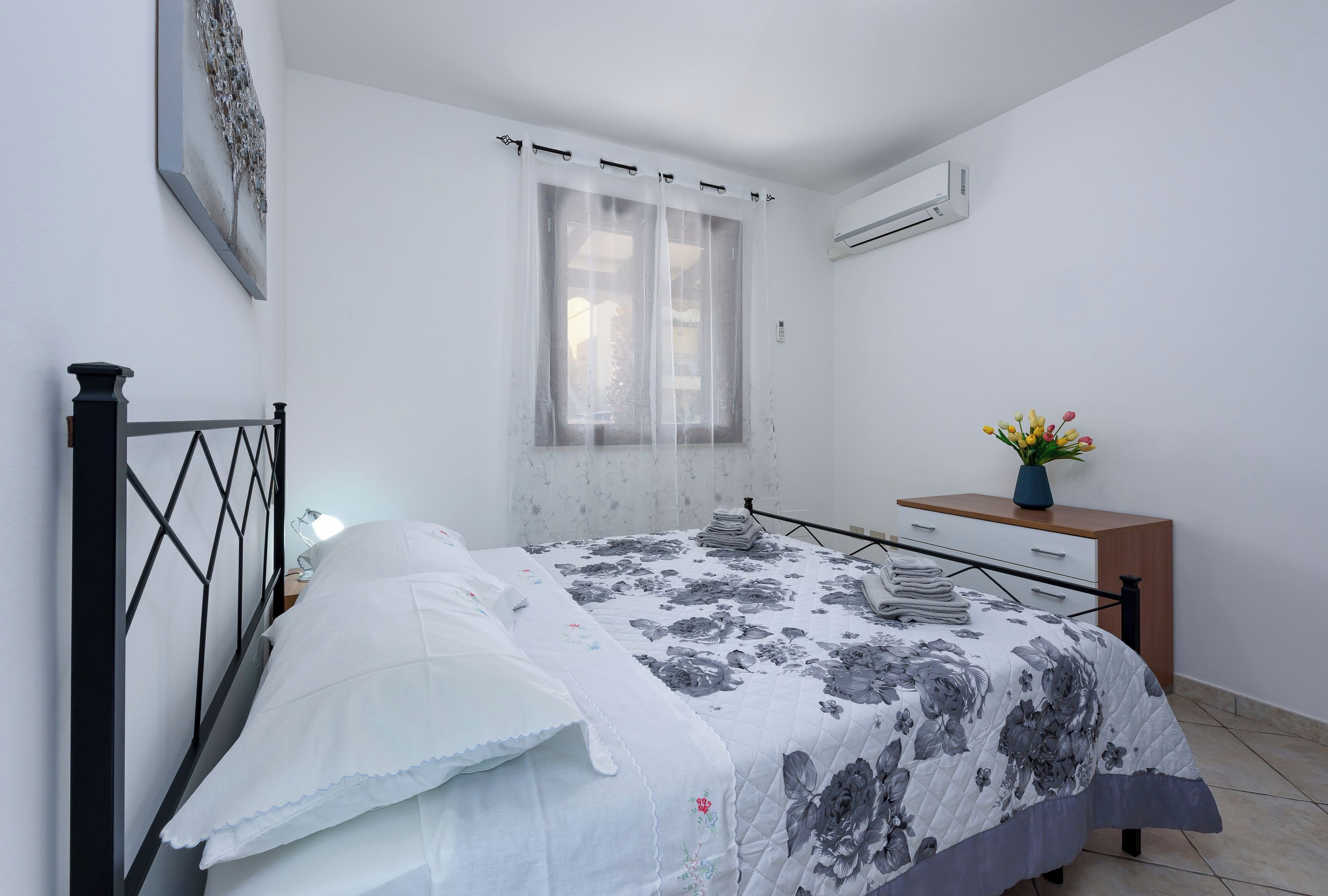 A bedroom with a bed, a nightstand, a dresser, a window with sheer curtains, and a wall-mounted air conditioner.