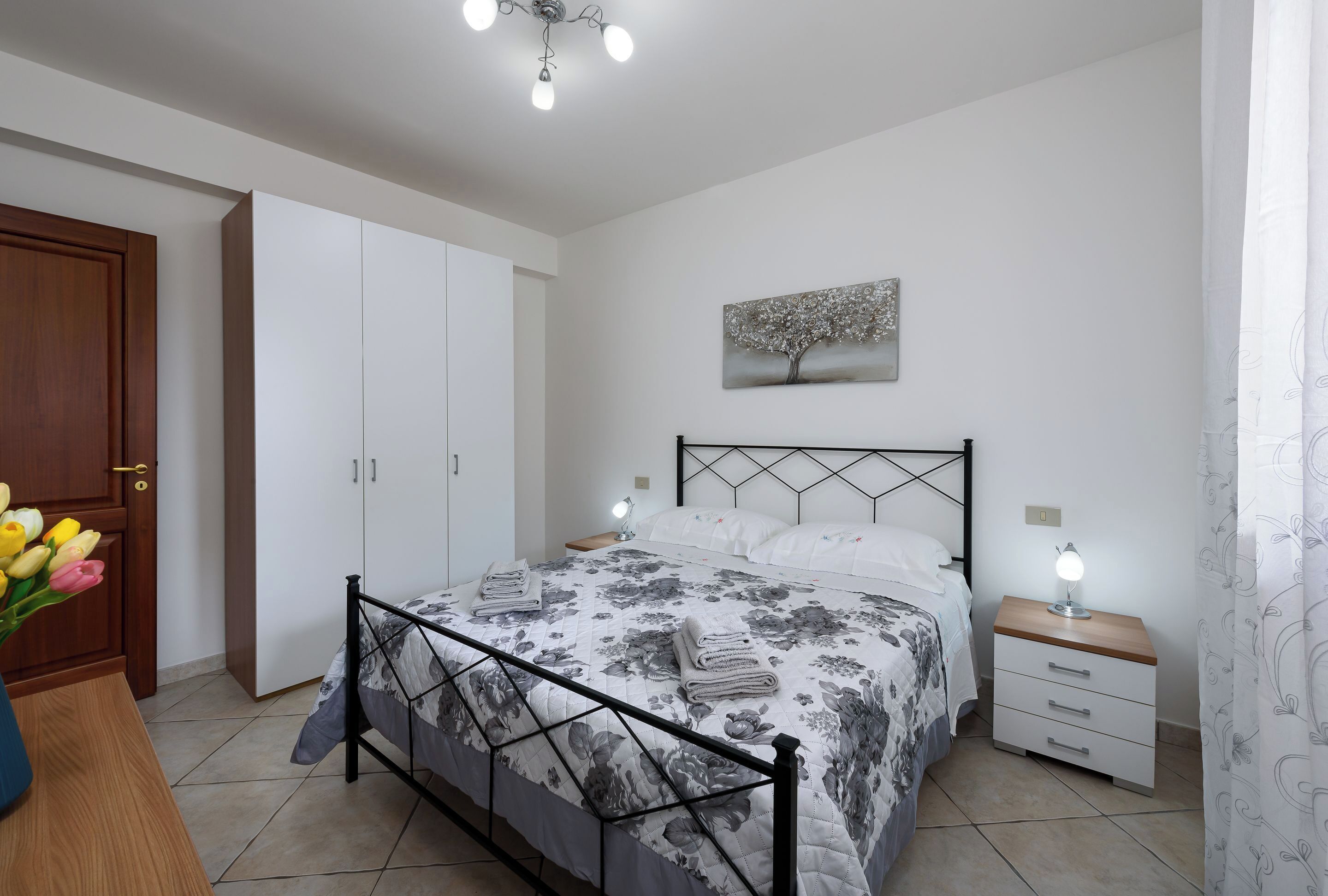 A bedroom with a bed, bedside tables, a nightstand, a wardrobe, a painting on the wall, and a vase of flowers.