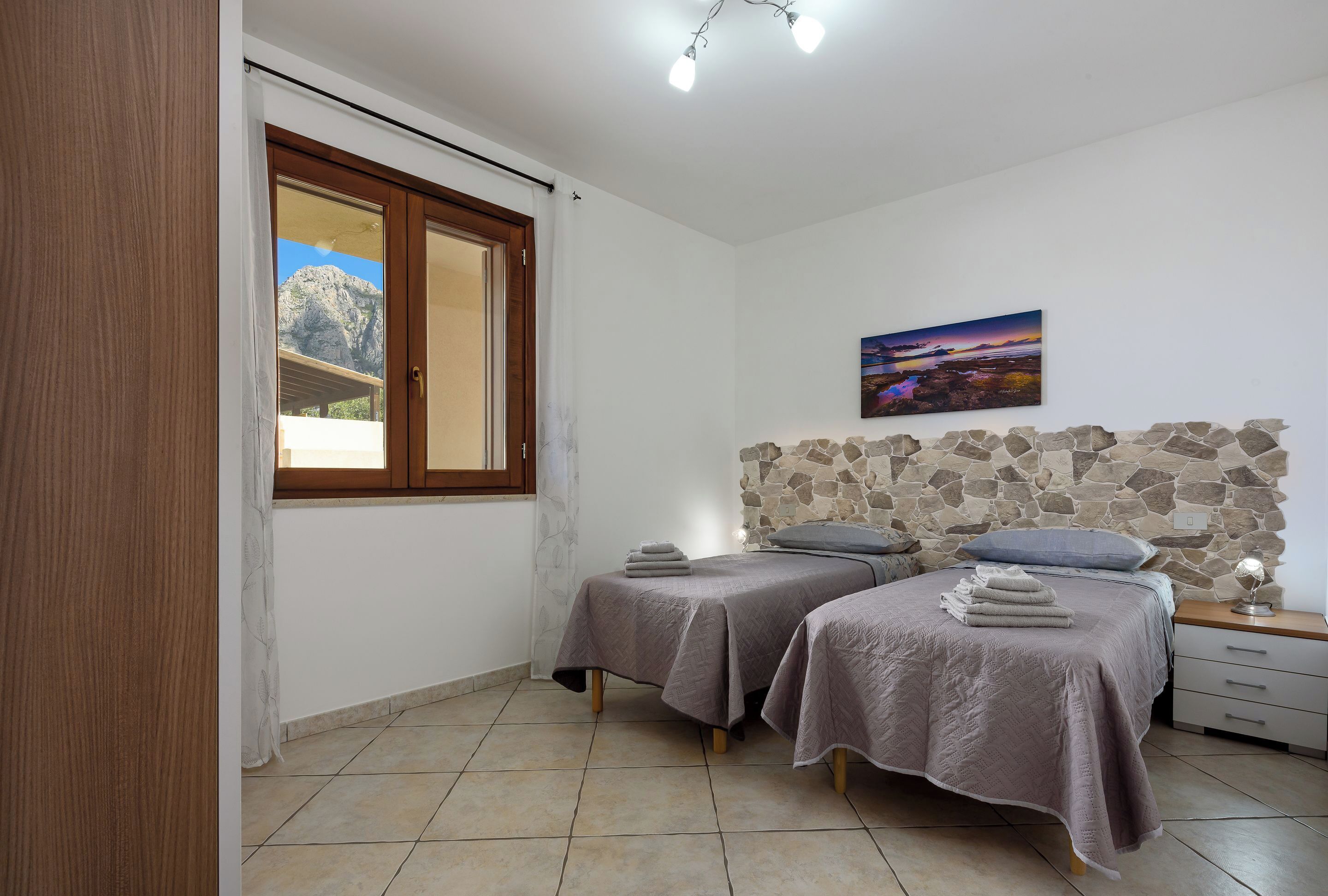 Un dormitorio con dos camas, una pared de piedra, un cuadro y una ventana con vistas a montañas.