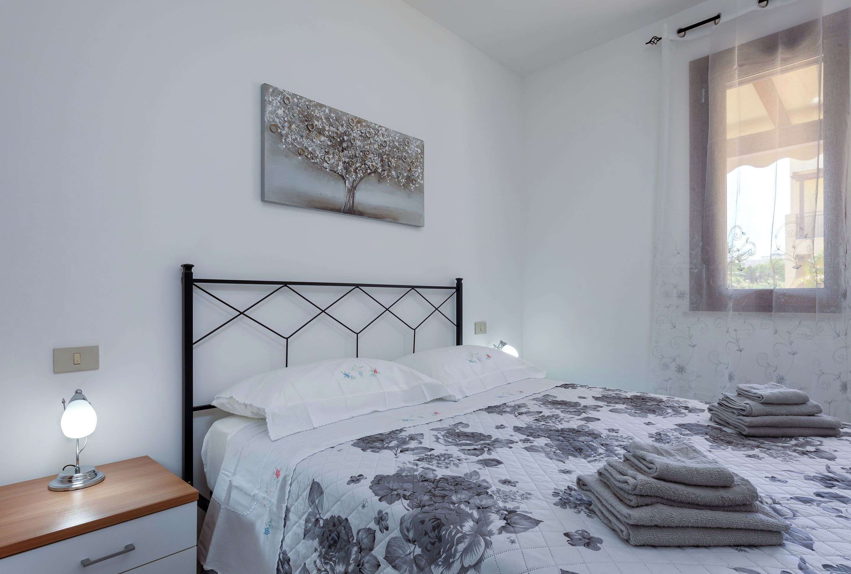 A bedroom with a bed, a nightstand with a lamp, a wall-mounted painting, and a window with curtains.