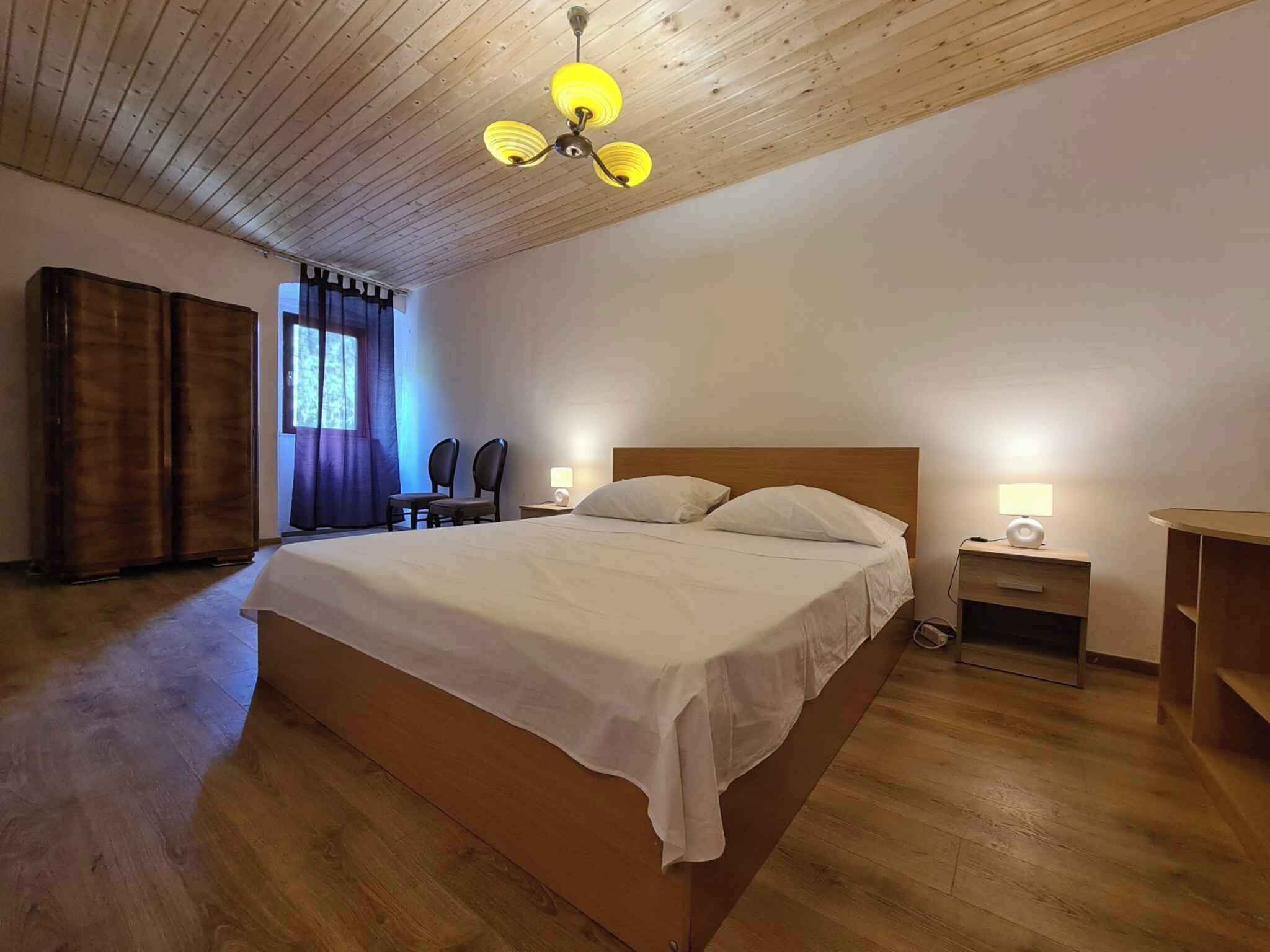 A bedroom with a wooden ceiling, a bed with white linens, two bedside tables with lamps, and a wooden wardrobe.