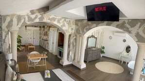 Quarto de hotel com uma cama grande, uma escrivaninha com cadeira, toucador com espelho e televisão fixada na parede.