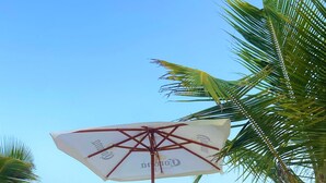 Cena de praia com duas cadeiras de praia brancas, uma mesa com frutas e um grande guarda-sol.