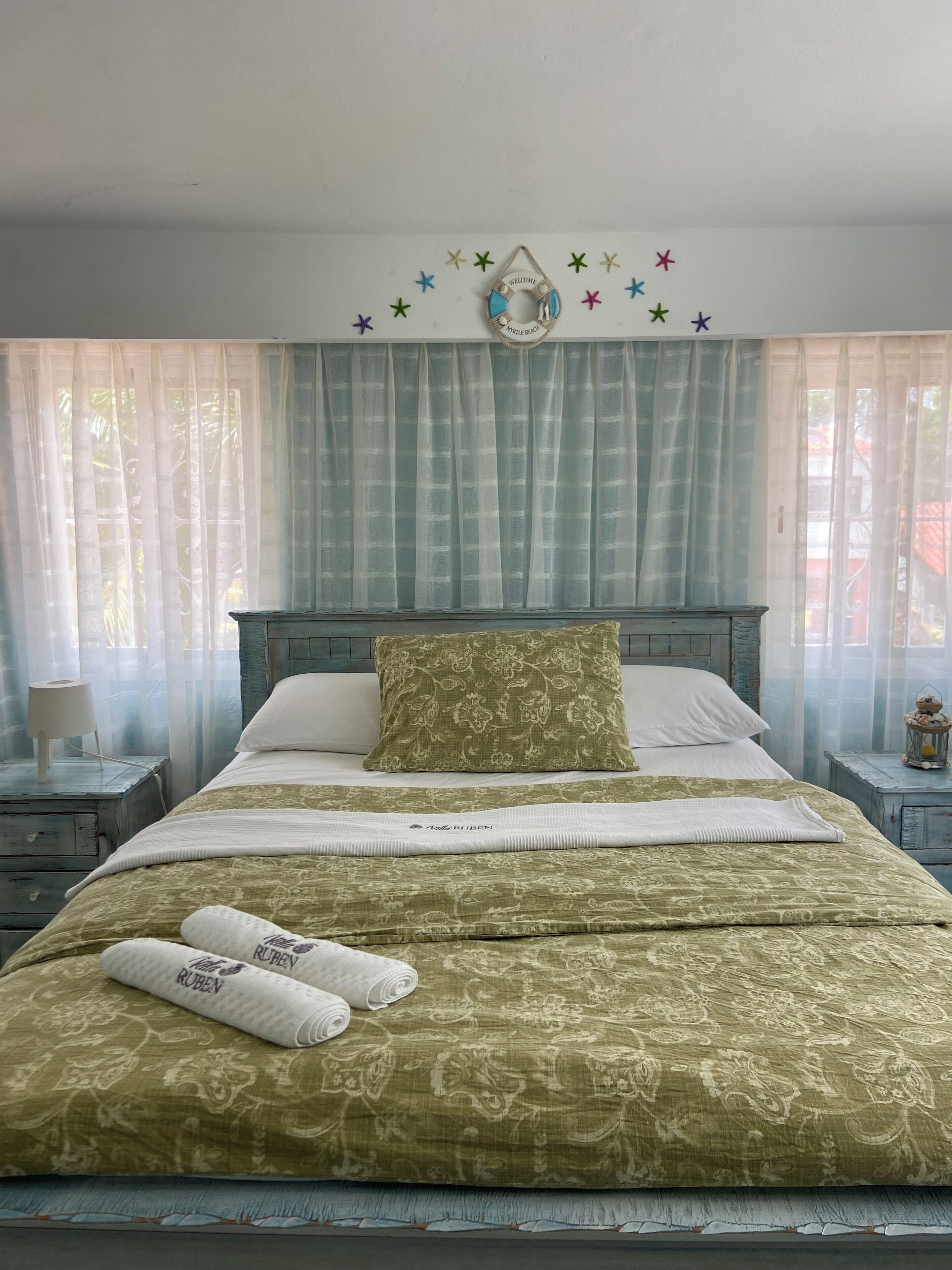 Un lit bien fait, recouvert d’une couvre-lit à motifs verts assortie aux coussins, une lampe de chevet et une décoration murale représentant des étoiles et un panneau d’accueil.