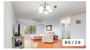 A spacious living room with a white ceiling, a chandelier, a ceiling fan, a large window, a white sofa, a patterned ottoman, and a small table. - Charming 2-bedroom condo with AC, WiFi in quaint Stuart (Stuart)