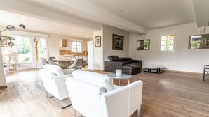 Amplia sala de estar con un moderno juego de sofá blanco, un sofá de cuero oscuro y una pequeña mesa redonda. Varias ventanas proporcionan luz natural, y se observa un área de cocina al fondo.