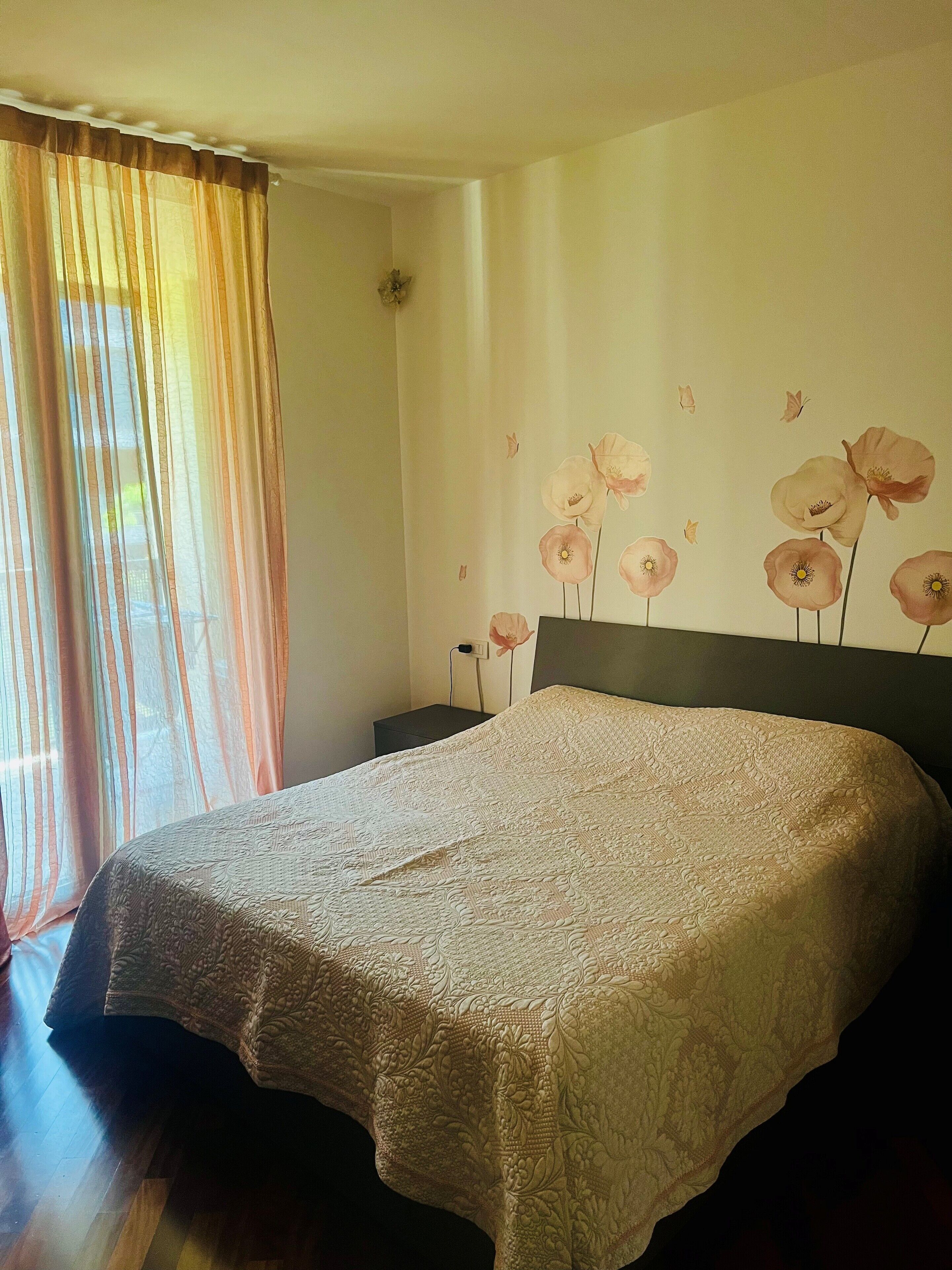 A bedroom with a large bed, a nightstand, a wall-mounted lamp, and a window with sheer curtains.