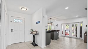 A modern kitchen with a central island, a dining area with a red table, and a white door with a stained glass window. - Orchard Oasis Retreat (West Palm Beach)