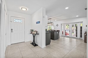 A modern kitchen with a central island, a dining area with a red table, and a white door with a stained glass window. - Orchard Oasis Retreat (West Palm Beach)