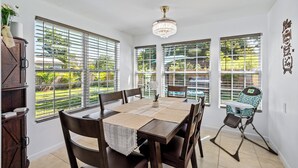 A dining area with a wooden table, chairs, and a high chair. Large windows with blinds allow natural light and views of greenery outside. - Orchard Oasis Retreat (West Palm Beach)