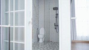 Une petite salle de bain fermée, équipée de toilettes et d’une douche, avec des murs et un sol carrelés, et une porte donnant sur une autre pièce.