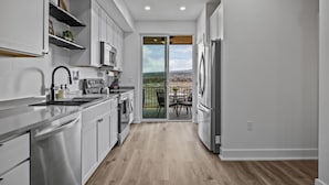 A modern kitchen with white cabinets, a stainless steel sink, and a built-in oven. There is a sliding glass door leading to a balcony with a view of the landscape.