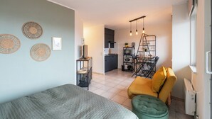 A compact living space with a bed, a green ottoman, a yellow chair, and a kitchenette in the background. - Studio in Blankenberge Near the Beach (Blankenberge)