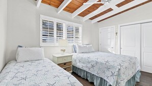A bedroom with two beds, a nightstand, a window with shutters, a ceiling fan, and a door.
