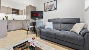 En moderne stue med en grå sofa, et sofabord med blomster, et fjernsyn der viser 'NETFLIX' og et køkkenområde i baggrunden.
