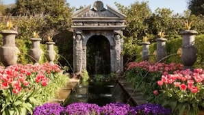 A garden with a central stone archway, surrounded by flower beds with red and purple flowers. - Arundel Cottage Retreat (Arundel)