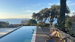 An infinity pool with a wooden deck and two lounge chairs, overlooking a body of water and hills. - Magnificent villa with panoramic view of the Mediterranean Sea (Le Lavandou)