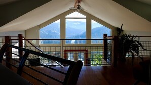 Una habitación con techo inclinado, ventanas amplias, vista a montañas, ventilador de techo y una escalera.