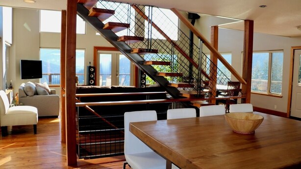 Un amplio salón con una escalera de madera, una mesa de comedor con sillas y un ventanal con vistas al exterior.