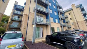 Un immeuble résidentiel à plusieurs étages, doté de balcons, d’un espace de stationnement et d’une voiture garée devant.