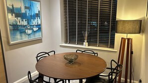 Un coin repas avec une table ronde en bois, des chaises noires, un tableau, une fenêtre avec des stores et un lampadaire.