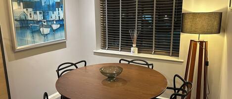 Un comedor con una mesa redonda de madera, sillas negras, un cuadro, una ventana con persianas y una lámpara de pie.