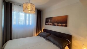 Een slaapkamer met een bed, een hanglamp, een schilderij aan de muur en een raam met gordijnen.