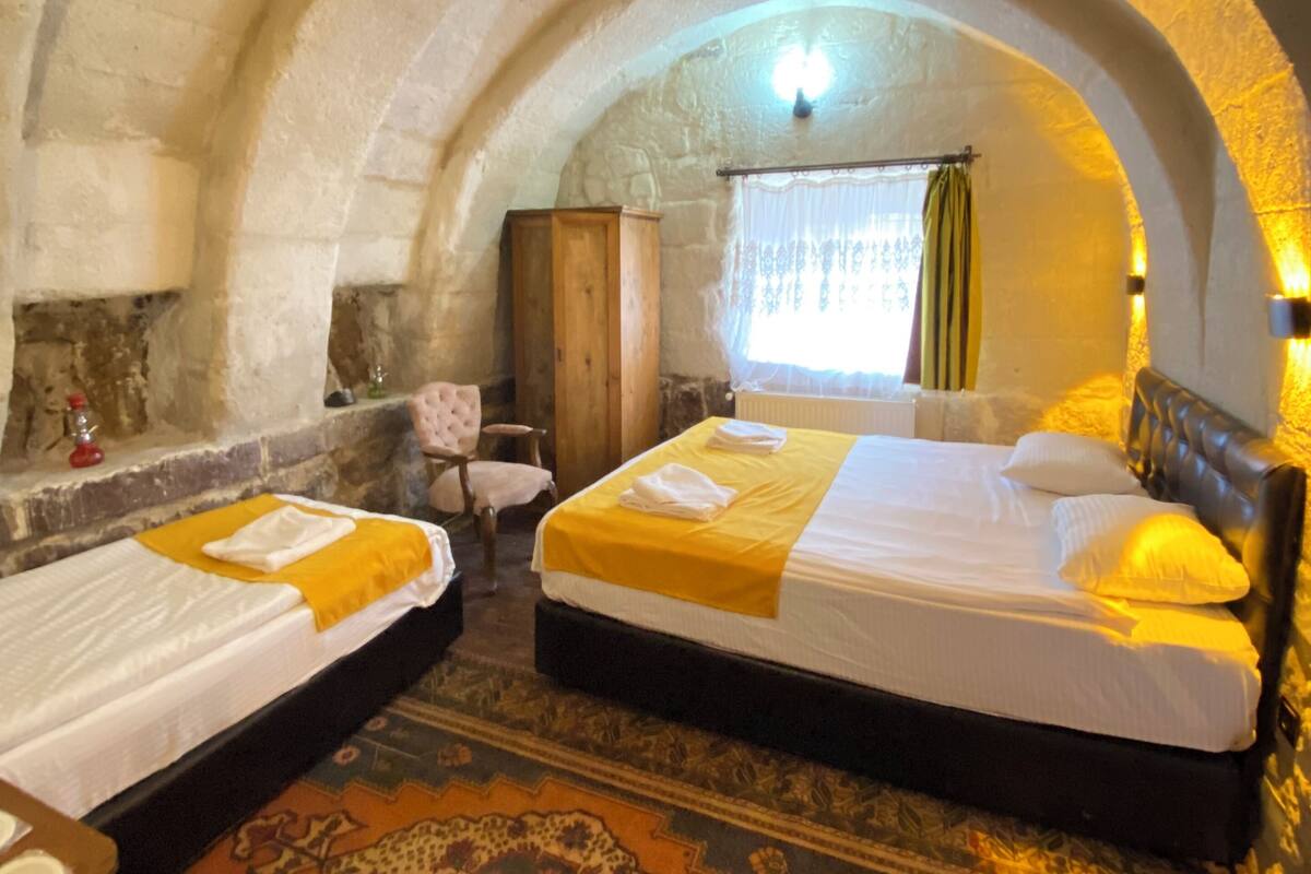 A room with two beds, a wooden wardrobe, a chair, a patterned rug, and stone walls.