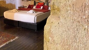 A room with a stone wall, a bed with red pillows, and a small table. - 7 Oda Kapadokya Cave House (Urgup)