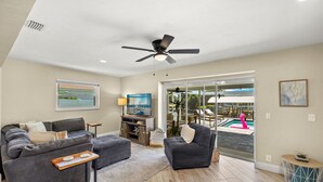 A spacious living room with a gray sofa, a small table, and a ceiling fan. A large window offers a view of a swimming pool and outdoor furniture.