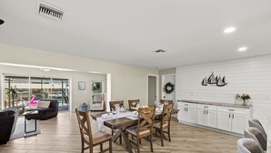 A spacious living area with a dining table set for six, wooden flooring, and a view of the outdoors through large windows.