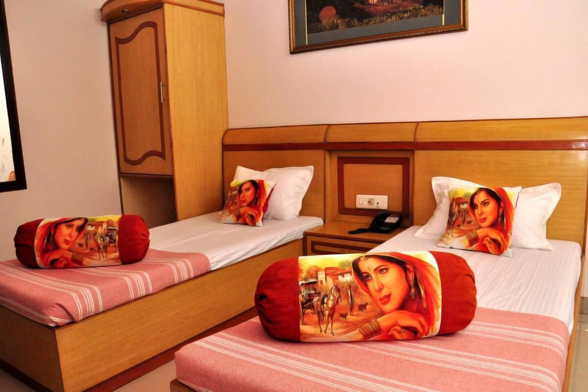 Habitación de hotel con dos camas individuales, cada una con un cojín que muestra el retrato de una mujer. Se observa un armario de madera y un cuadro en la pared sobre las camas.