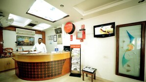 A reception area with a wooden reception desk, a television mounted on the wall, and a framed Buddha statue. - Hotel Blue Bell (New Delhi)