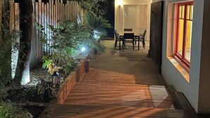 Un sendero de madera conduce a un área de comedor con una mesa y sillas, rodeado por una cerca y árboles.