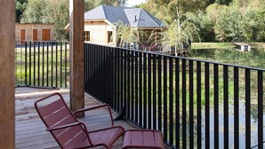 A wooden deck with red metal chairs, a black metal railing, and a view of a house and trees across a body of water. - Escape to Nature in Vencimont (Gedinne)