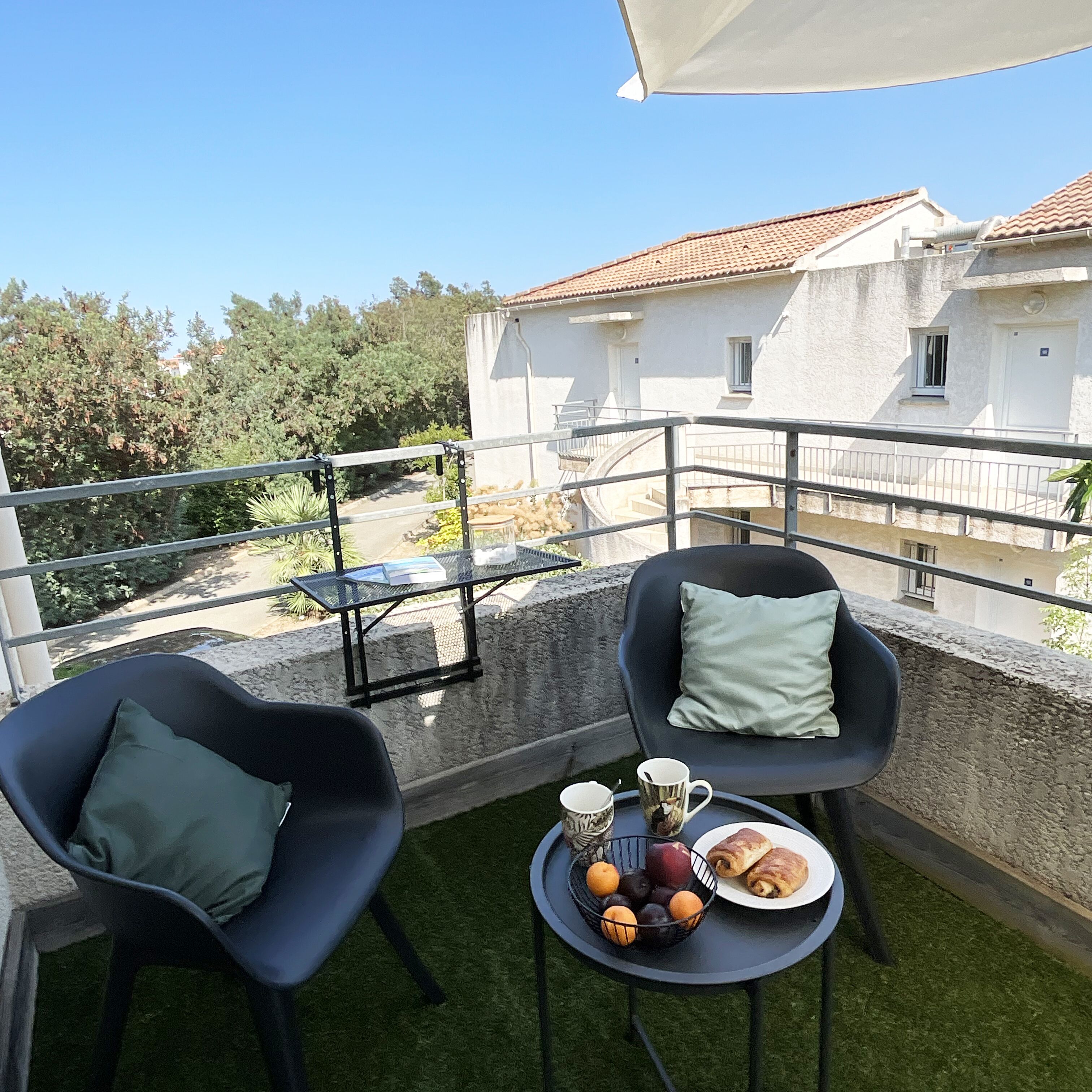 A balcony with two black chairs, a small round table with a fruit bowl, two mugs, and a plate of pastries.