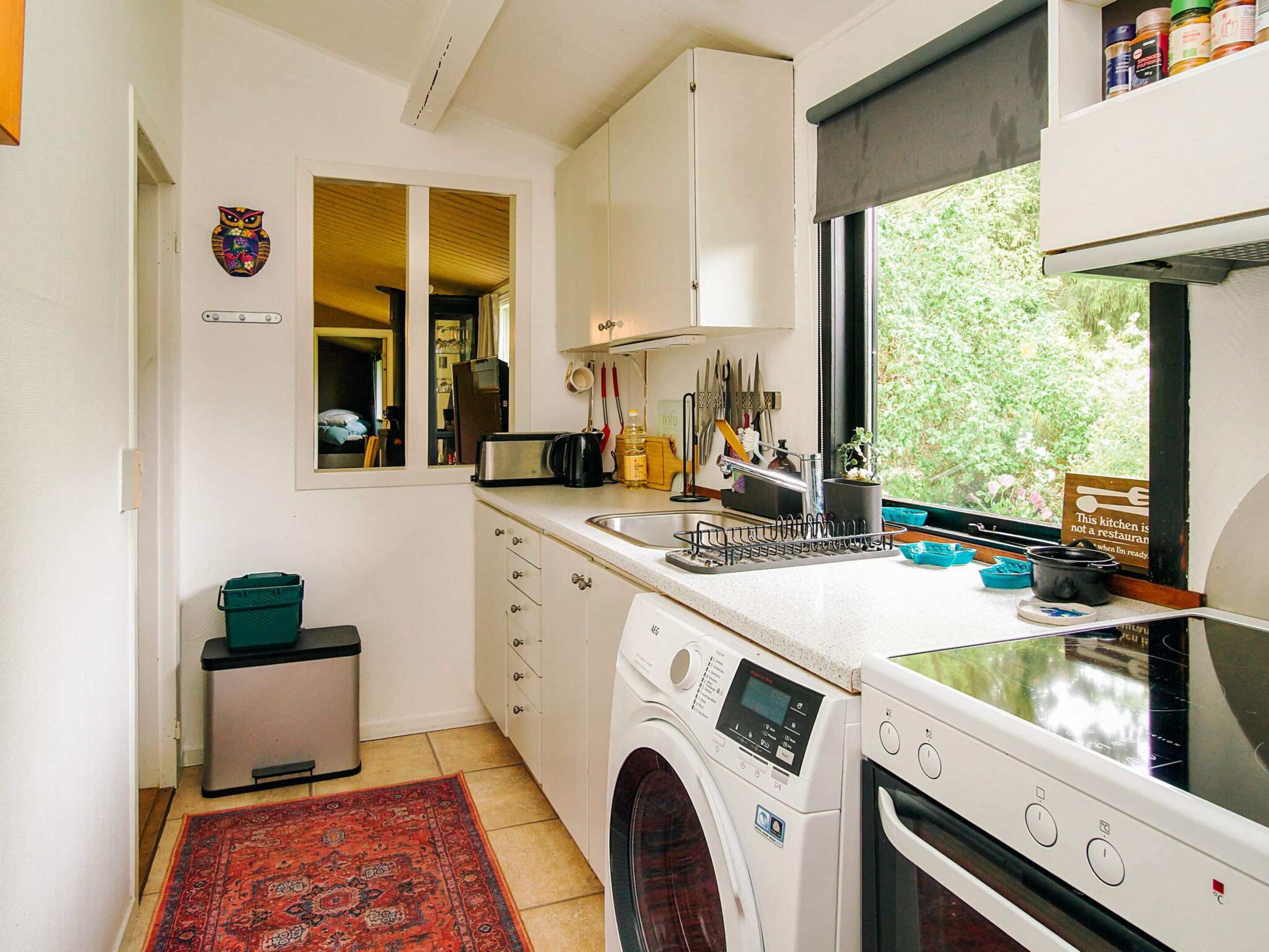 A compact kitchen with a washing machine, a small refrigerator, and a window with a view of greenery.