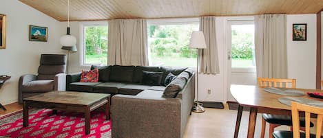 A cozy living room with a sofa, armchair, coffee table, and dining table. There are windows with curtains, a lamp, and artwork on the walls.