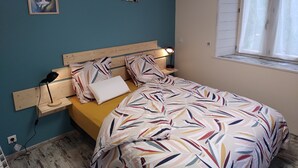Une chambre à coucher avec un lit, une table de chevet, une lampe, un tableau et une fenêtre avec des rideaux.