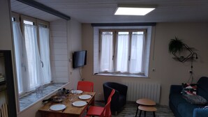 Un coin repas avec une table en bois dressée pour quatre personnes, des chaises rouges, un canapé noir et un divan bleu. Un téléviseur est fixé au mur, et une fenêtre avec des rideaux est visible.
