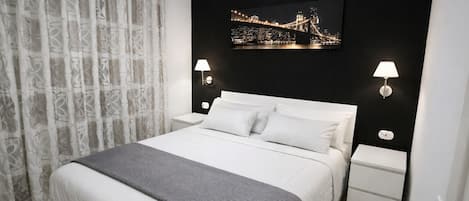 A hotel room with a bed, two bedside tables, a nightstand, a gray throw blanket, a black wall with a framed picture, and a white curtain.